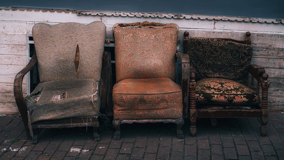 The image displays three vintage upholstered armchairs placed outdoors on a brick-paved surface against a weathered white wooden wall with a corrugated metal roof visible at the top. The chair on the left features a light brown, floral-patterned fabric that is severely torn, revealing foam padding inside and frayed edges along the seat and armrests. The middle chair has an aged, orange-tan fabric with a floral motif, appearing worn and slightly faded, with visible stains and a sagging seat cushion. The rightmost chair is a wooden-framed armchair with intricately carved details and a patterned fabric cushion in shades of black and gold, which appears less damaged but still aged. The scene suggests these chairs may be intended for private collection, disposal, or awaiting removal by a waste clearance service such as Rubbish Clearance Bow, reflecting an informal collection of furniture set rather than a curated display, emphasizing the importance of professional rubbish removal for such items.