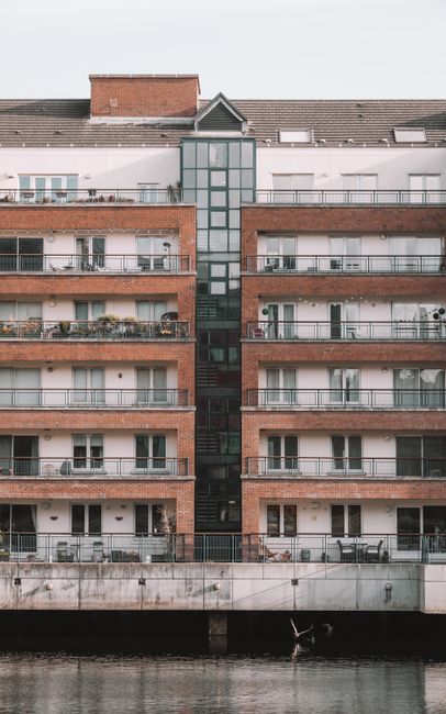 A multi-story residential building with a brick facade and numerous balconies featuring metal railings, visible outdoor furniture, and plants. The building has a central glass stairwell with dark framing extending vertically through the middle, connecting the floors. The structure is situated beside a waterway, with a concrete quay or promenade at the base, and the water reflects parts of the building. The sky overhead is overcast, creating diffuse natural lighting that highlights the textures of the brickwork and glass. The overall scene reflects an urban riverside environment, where private apartments overlook the water, and the setting could be associated with general property management and waste disposal challenges typical of riverside housing areas. Rubbish Clearance Bow might handle waste removal for such homes through external collection services, as indicated by the urban setting and external environment, without any visible waste or removal equipment in the image.